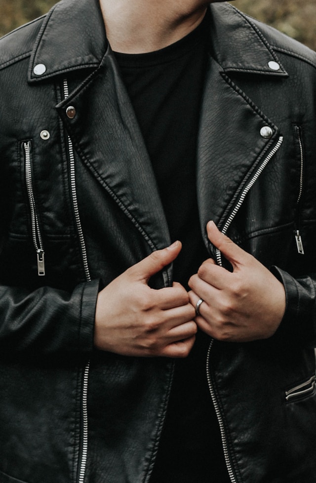 A man in a black leather jacket stands confidently with his hands in his pockets against a backdrop of vibrant green foliage. He has short, curly hair and is wearing round glasses. The image captures a casual, yet stylish look, with the greenery adding a refreshing contrast to his dark outfit.