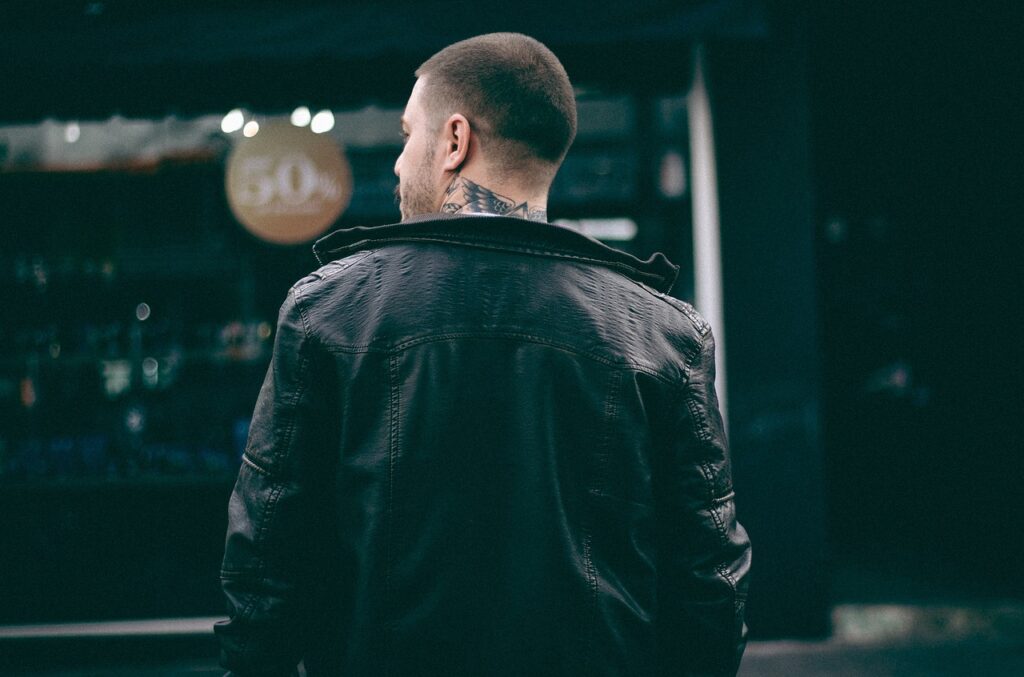 A man in a dark brown leather jacket stands on a city street with a contemplative expression. He has short, dark hair and a trimmed beard. The background features a blurred view of urban buildings and pedestrians, suggesting a bustling city environment.